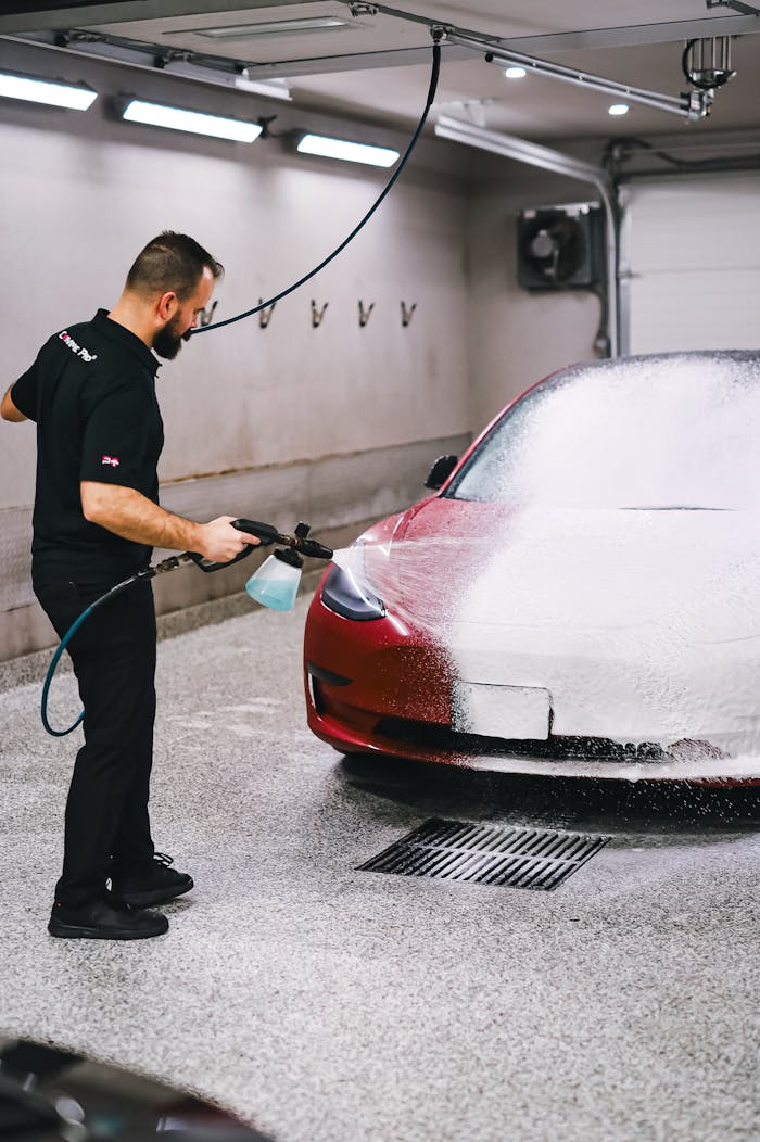 Man using high-pressure hose to wash a red car in an indoor garage setting with foam.